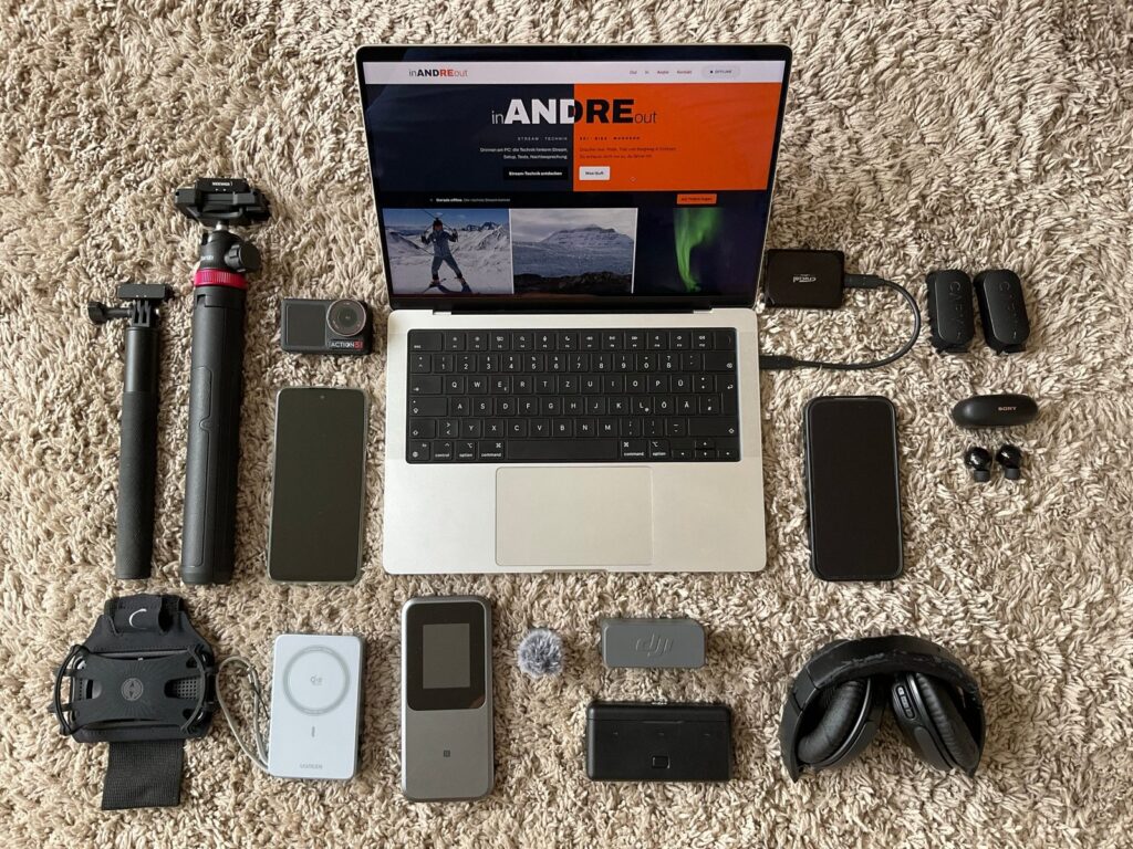 Home Flatlay des mobilen Stream-Setups: MacBook Pro mit der Seite inANDREout, iPhone, Motorola, Action 5 Pro, Mikrofone, Router, Powerbank, Stative und Kopfhörer